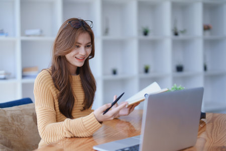 Smiling young woman engaged in a joyful online e-learning video call, gesturing while holding a book with headphones on a wooden table nearby.の写真素材