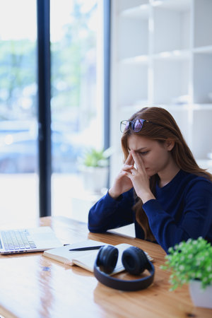 Stressed young woman overwhelmed by e-learning studies at desk with laptop, notebook, headphones, hands clasped to her face in frustration.の写真素材