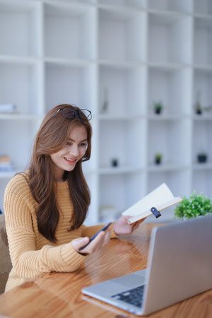 Smiling young woman engaged in a joyful online e-learning video call, gesturing while holding a book with headphones on a wooden table nearby.の写真素材