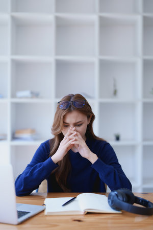 Stressed young woman overwhelmed by e-learning studies at desk with laptop, notebook, headphones, hands clasped to her face in frustration.の写真素材