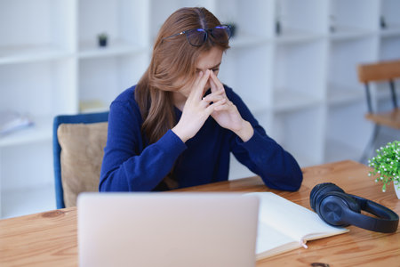Stressed young woman overwhelmed by e-learning studies at desk with laptop, notebook, headphones, hands clasped to her face in frustration.の写真素材