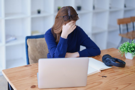 Stressed young woman overwhelmed by e-learning studies at desk with laptop, notebook, headphones, hands clasped to her face in frustration.の写真素材