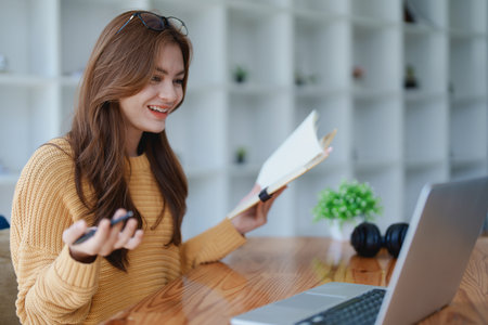 Smiling young woman engaged in a joyful online e-learning video call, gesturing while holding a book with headphones on a wooden table nearby.の写真素材