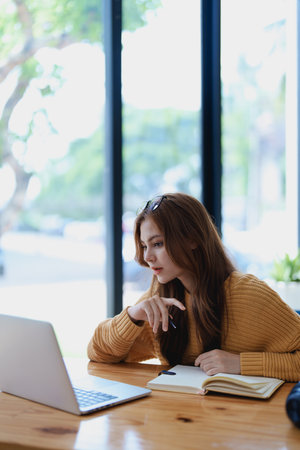 Smiling young woman engaged in a joyful online e-learning video call, gesturing while holding a book with headphones on a wooden table nearby.の写真素材