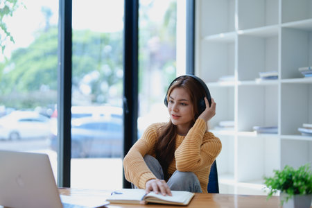 Young woman enjoying a relaxing music break at her desk while using headphones, writing in a notebook, and working on her laptop for calm inspiration.の写真素材