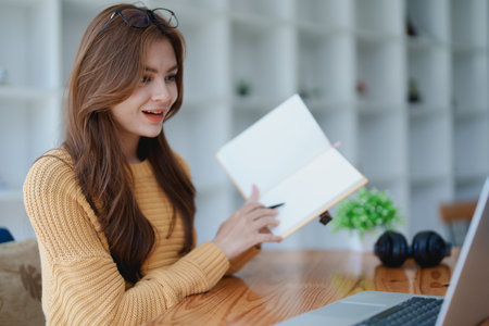 Smiling young woman engaged in a joyful online e-learning video call, gesturing while holding a book with headphones on a wooden table nearby.の写真素材