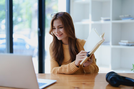 Smiling young woman engaged in a joyful online e-learning video call, gesturing while holding a book with headphones on a wooden table nearby.の写真素材