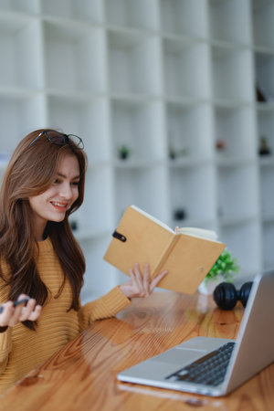 Smiling young woman engaged in a joyful online e-learning video call, gesturing while holding a book with headphones on a wooden table nearby.の写真素材