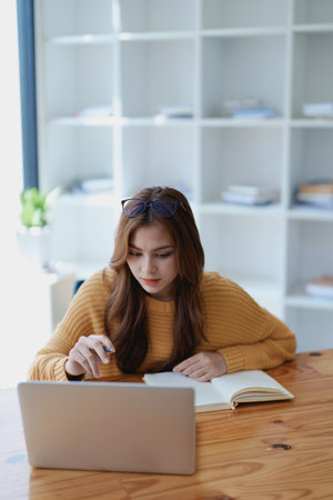 Smiling young woman engaged in an online e-learning video call, gesturing while holding a book with headphones on a wooden table nearby.の写真素材