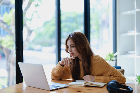 Smiling young woman engaged in a joyful online e-learning video call, gesturing while holding a book with headphones on a wooden table nearby.の写真素材
