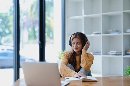 Young woman enjoying a relaxing music break at her desk while using headphones, writing in a notebook, and working on her laptop for calm inspiration.の写真素材