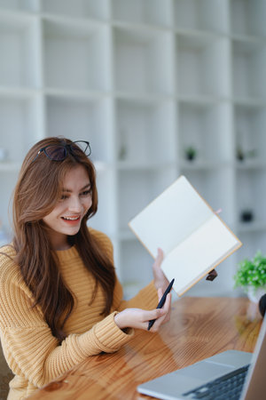 Smiling young woman engaged in an online e-learning video call, gesturing while holding a book with headphones on a wooden table nearby.の写真素材