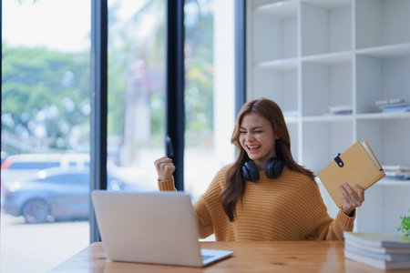 Young woman celebrates success with laptop and book at her bright workspace.の写真素材