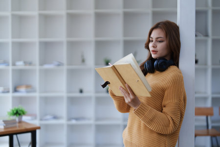 Young woman reads a book with headphones around her neck in a bright room..