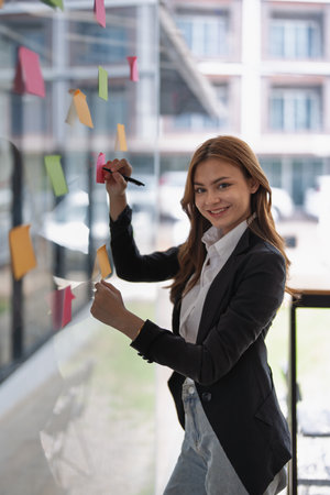 An asian businesswoman uses sticky notes on glass in a modern office space..の写真素材