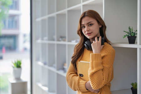 Young pretty woman leans on bookshelf holding a book and headphones..の写真素材