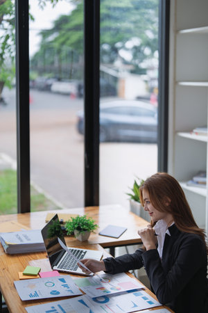 Young businesswoman analyzes charts and graphs on laptop in bright office.の写真素材