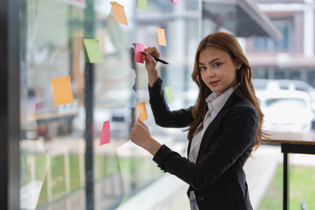 Businesswoman writing ideas on sticky notes on glass wall in office space..の写真素材