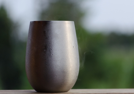 frosty beverage in silver stainless steel stemless wine glass cup outside with condensation drops and steam green trees behindの写真素材