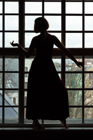 Silhouette of a woman in a red dress with head turned in front of a small pane windowの写真素材