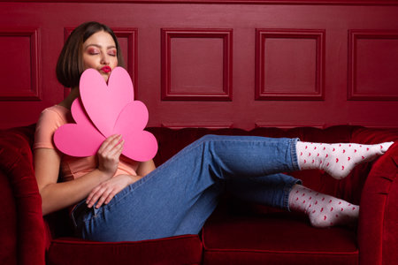 Women in jeans with closed eyes on a couch holding three heart-shaped paper sheets and lips shaping kissの写真素材
