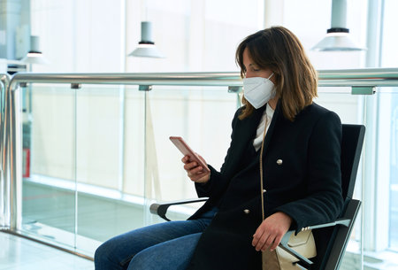 pretty brunette girl with protective mask looks at the mobile sitting in a station. copy spaceの写真素材