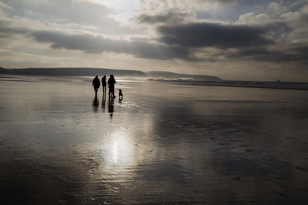 Monochromatic, backlit beach scene with people in the distance and reflections of the sky on the wet sand.の写真素材