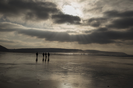 Monochromatic, backlit beach scene with people in the distance and reflections of the sky on the wet sand.の写真素材