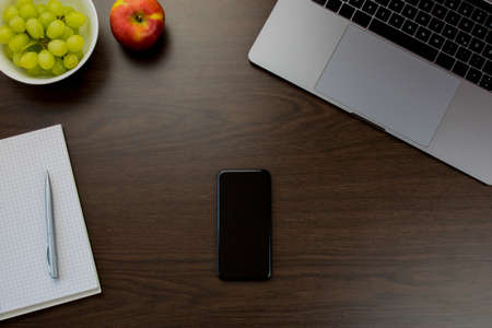 Desk in the office with a notebook next to grapes and an apple, a notepad and pen, a smartphone in the middle - wooden tableの写真素材
