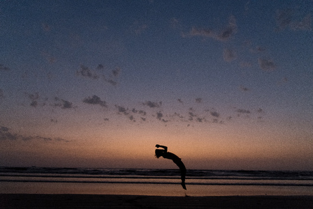 On a hot summer evening youth performs somersaults, flips  handstands at  beach. Silhouetted at dusk with clouds in  sky.の写真素材