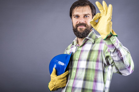 Happy young worker with hardhat and gloves showing ok sign over gray backgroundの写真素材
