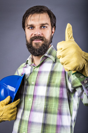 Happy young worker with hardhat and gloves showing ok sign over gray backgroundの写真素材