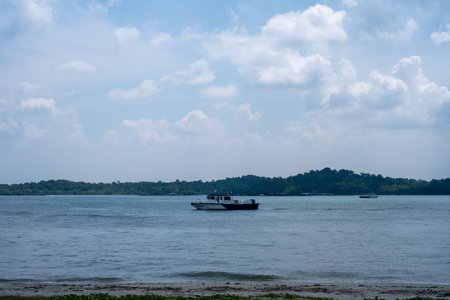 Small boat floats on calm blue water. Sky holds scattered white clouds above. Tree-lined shore forms...