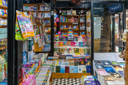 Colorful books line shelves in a cozy bookstore. Stacks of magazines sit on counters nearby. Brightly packaged items hang on display racks. Wooden shelves hold assorted children's literature