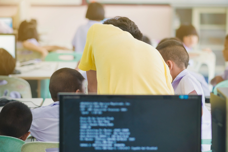 Male teacher in yellow shirt going around class, helping and checking works of pupils in computer classroom.の写真素材