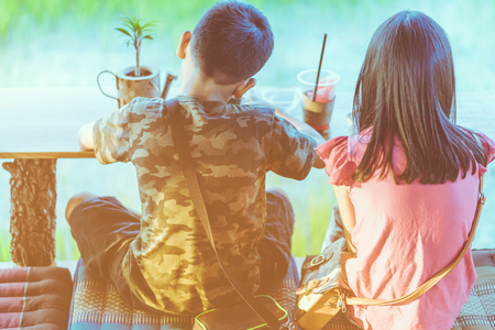 Back view of brother and sister are playing mobile phone games and relaxing with beverage at the cottage in the rice fields.の写真素材