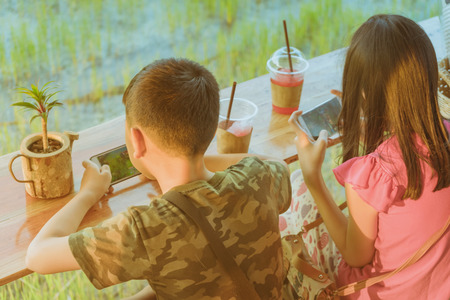 Back view of brother and sister are playing mobile phone games and relaxing with beverage at the cottage in the rice fields.の写真素材