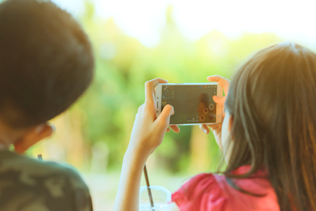 Back view of brother and sister are playing mobile phone games and relaxing with beverage at the cottage in the rice fields.の写真素材