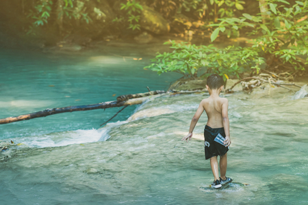 Little boy crossing a river full of stones carefully and teetering in waterfall at Erawan National Park in Kanvhanaburi,Thailand.の写真素材
