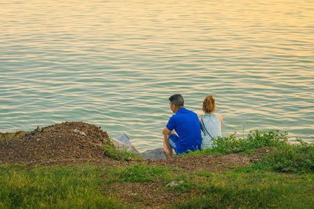 Back view of young couple sitting together on stone in front of ocean enjoying sunset during summer vacation.の写真素材