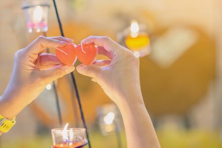 A red heart shaped candle in a woman's hand with candlelight in many small glass cups. A valentine's day background.の写真素材