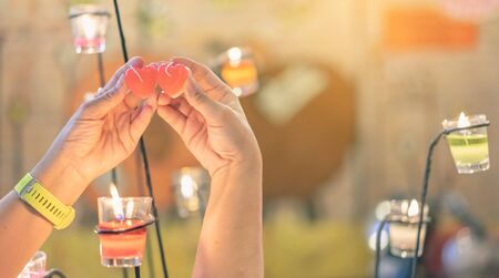 A red heart shaped candle in a woman's hand with candlelight in many small glass cups. A valentine's day background.の写真素材