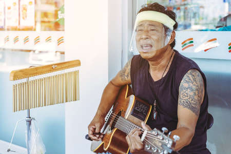 KANCHANABURI,THAILAND-APRIL 18,2020: Unidentified street musician wear a face mask playing guitar and tubular bells in front of 7-Eleven at Thamuang district on april 18,2020 in Kanchanaburi,Thailand.のeditorial素材