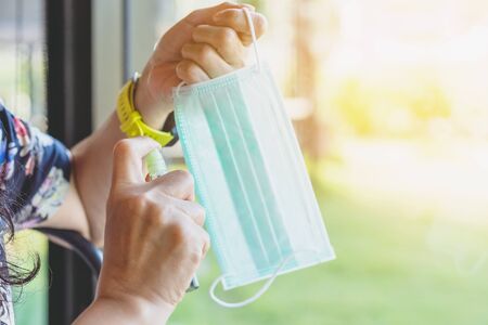 Hand of woman is spraying alcohol on old face mask to prevent infection of corona virus (Covid-19)...