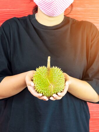 Young woman wear a protective mask to prevent infection of corona virus (Covid-19) in a black shirt holding a ripe durian, ready to eat with the red wooden wall. New normal life style concept.の写真素材