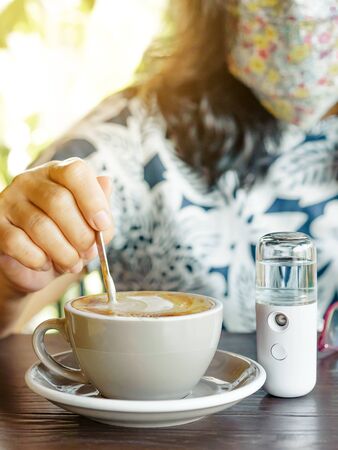 A quarantined woman relax with hot cappuccino coffee and alcohol nano mist sprayer on table while being quarantined due to the Corona virus (Covid-19) outbreak. New normal concept. Selective focusの写真素材