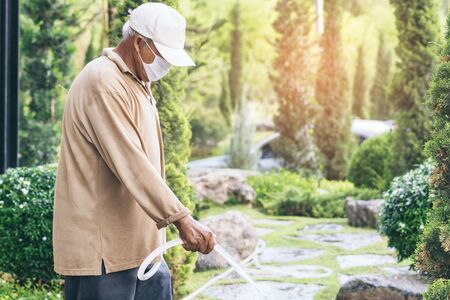 A quarantined elderly man wear a face mask to prevent the spread of the Corona virus (Covid-19) watering plants for exercise in the garden at home. New normal lifestyle.の写真素材