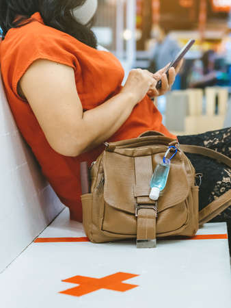 Mini alcohol gel bottle to kill Corona Virus(Covid-19) hang on a leather shoulder bag of a woman wearing a mask while use smartphone with alternative seating mark for social distancing in the mall.の写真素材
