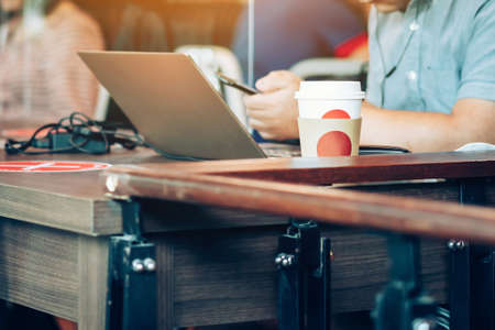 Man work on computer with smartphone and relax with hot coffee in coffee shop, where seats are spaced for social distancing during the Corona virus (COVID-19) epidemic. Selective focus on coffee cup.の写真素材
