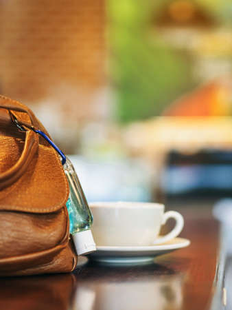 Mini portable alcohol gel bottle to kill Corona Virus(Covid-19) hang on a brown leather shoulder bag on table in garden. New normal lifestyle. Health care concept. Selective focus on alcohol gelの写真素材